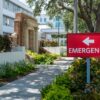 A pleasant sidewalk under a canopy of trees outside a hospital, with a sign pointing to the Emergency department