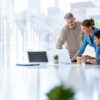 Three colleagues collaborate around a laptop in a bright, airy office conference room